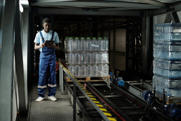 Fototapeta na wymiar Young African American factory worker in coveralls using tablet while standing by conveyor belt with capped plastic bottles with drinks