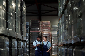 Fototapeta na wymiar Two young factory workers in coveralls standing among stacks of packages with bottled water and discussing data on tablet screen