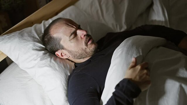A bald hispanic man with a beard appears in discomfort lying in bed in a dark bedroom at night, symbolizing insomnia or illness.