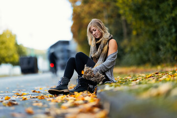 A young blonde woman in a knitted earth-colored look with black leggings and blue boots sits on a sidewalk covered in autumn leaves.