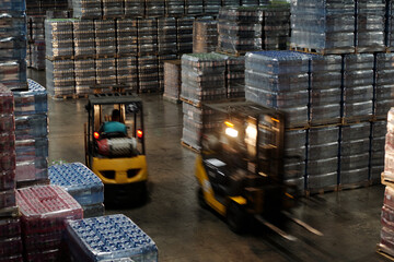 Blurry motion of two forklifts along aisle between huge stacks of packed plastic bottles containing...