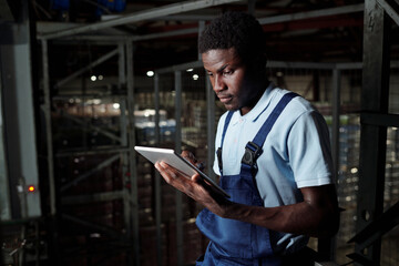 Young male engineer of factory warehouse with tablet scrolling through data on screen while standing by racks with packages