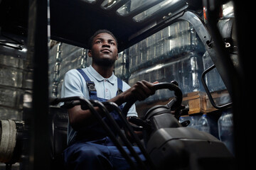 Young worker of huge industrial warehouse in coveralls sitting in forklift and driving it while...