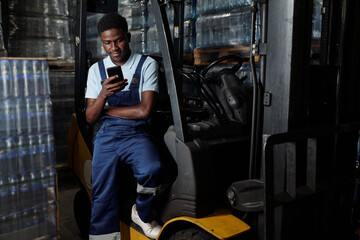 Fototapeta na wymiar Young male engineer of industrial plant sitting by door of forklift and texting in mobile phone while having break after work