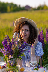 Beautiful happy young woman on a meadow arranging table for outdoor event, gathering wildflowers, lighting candles. Wedding or romantic date decoration in the field with purple lupins, fruits