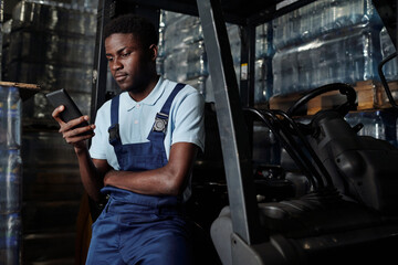 Fototapeta na wymiar Young factory engineer sitting against door of forklift at break and texting in cellphone in distribution warehouse with stacks of bottled water