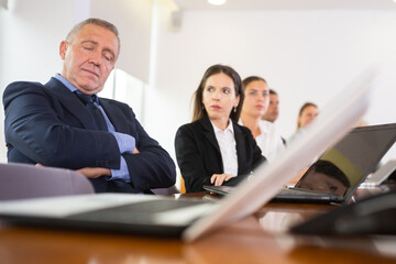 Exhausted mature white office employee in formal suit having a nap during business meeting in conference room with his arms crossed
