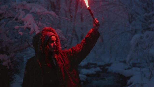 Portrait: A young girl holding a burning signal fire. A woman in a winter forest lights a red rescue flare. Snow on the trees, blue hour. Female person gives a distress signal. Snowy weather
