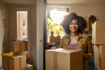 Smiling afro-american woman removes to her new home. Portrait of curly woman with cardboard boxes. Flower pots with plants, folded clothes and other things.  - 728850218