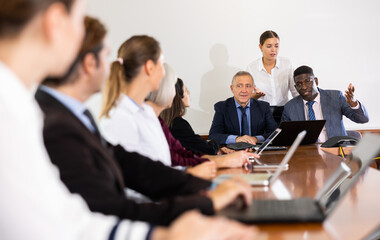 Different aged multiethnic company workers in formal wear sitting at table in meeting room during briefing.