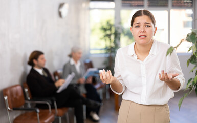 Portrait of worried upset young female employee standing in office lobby with papers in hands..