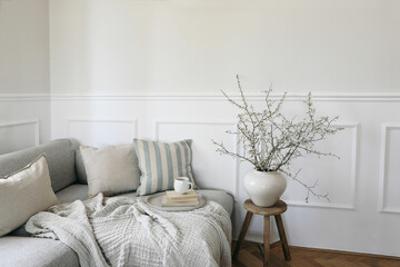 Modern spring scandinavian living room interior. Sofa with linen pale blue striped cushions and cup of coffee. Cherry plum blossoms in vase. Elegant stylish minimal home decor. Easter breakfast.