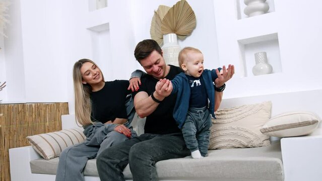 Caucasian parents with infant baby sit on the sofa. Dad supports his tiny son who's starting to walk.