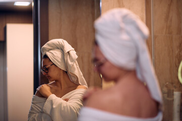 A woman gracefully applies nourishing moisturizer to her shoulder after a relaxing bath, embodying...