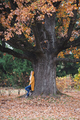 A beautiful blonde woman is standing by the wide old oak tree in autumn. 