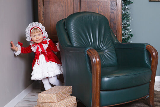 A little girl in a festive red velure dress and hat by the green chair