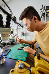 side view of asian technician in goggles making technical diagnostics of smartphone in repair shop