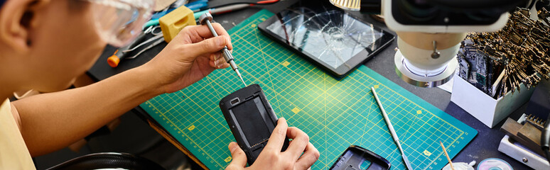 cropped view of repairman in goggles with screwdriver fixing mobile phone at workplace, banner