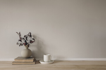 Breakfast still life. Neutral Scandinavian interior. Minimal home design. Ceramic vase with dry eucalyptus tree branches. Cup of coffee on wooden table, desk with old books. Empty beige wall mockup.