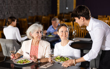 Smiling waiter fulfills order of young girl and elderly woman visiting restaurant. Women customers of cafe happy to have pleasant service of small family cafe