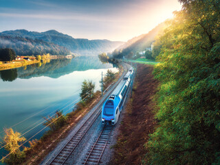 Aerial view of blue modern high speed train moving near river in alpine mountains at sunrise in summer. Top view of train, railroad, lake, reflection, trees in spring. Railway station in Slovenia - obrazy, fototapety, plakaty