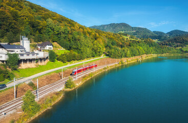 Fototapeta premium Aerial view of red modern high speed train moving near river in alpine mountains at sunny spring day. Top view of train, rural railroad, lake, road, green trees in summer. Railway station in Slovenia