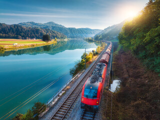 Aerial view of red modern high speed train moving near river in alpine mountains at sunrise in...