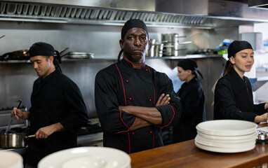 Focused man executive chef standing in modern restaurant kitchen, posing on camera, while his team...