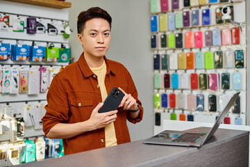 confident asian salesman with mobile phone looking at camera near laptop on counter in private shop