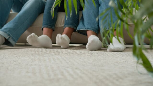 Moving shot of male female legs in socks on floor in living room diverse family mother mom father dad parents and son little boy kid relax on couch sofa at home adult and child feet underfloor heating