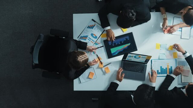 Top down aerial view of diverse business people discuss and brainstorm marketing idea while looking at financial graph at table with marketing document and sticky notes scatter around. Directorate.