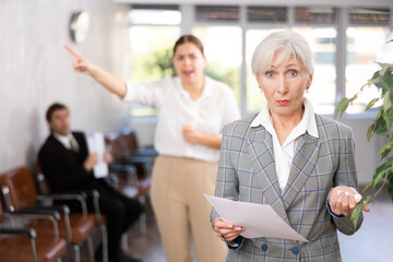 Upset elderly woman in business suit with documents in her hands being criticized in office