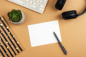 Desktop with blank card, notepad, computer keyboard, succulent plant, pen and headphones on brown...