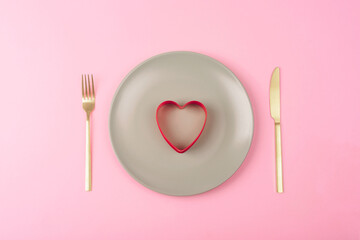 Gray plate with red cookie cutter in a shape of heart, fork and knife on pink background. Valentines day concept. Top view, flat lay