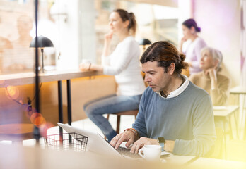 Obraz premium Concentrated male designer preparing new project on laptop, sitting in cozy cafe with cup of coffee