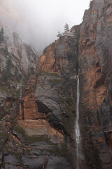 An ephemeral waterfall pours down the wet red and black sandstone cliffs of Zion Nat. Park in Utah, USA while low clouds fill the canyon above.