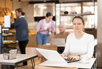 Fototapeta premium Focused woman drinking coffee while working on laptop in cozy cafe