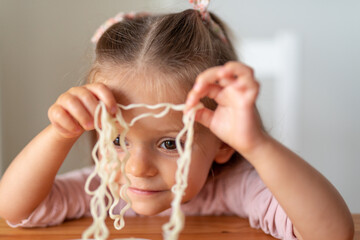 Cheerful little child toddler girl eating and playing with Asian noodles in a light white kitchen