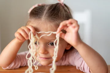 Zelfklevend Fotobehang Cheerful little child toddler girl eating and playing with Asian noodles in a light white kitchen © Razbitnov