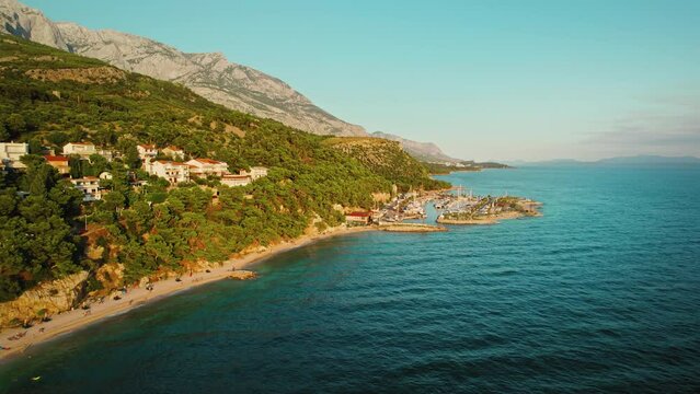 Seaside town and calm water of Adriatic Sea at golden hour. Coastal buildings on rugged slopes of Biokovo mountain in Croatia.