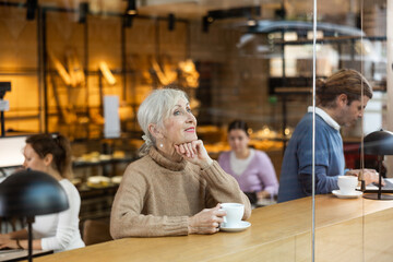 Smiling mature female in casual wear sitting at table and enjoying coffee or tea in cozy cafeteria