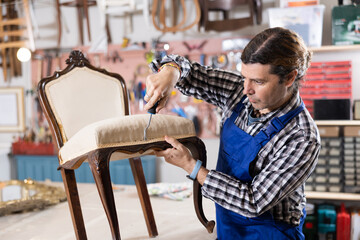 unhurried male restorer with hand tool removes old upholstery from vintage chair with twisted legs