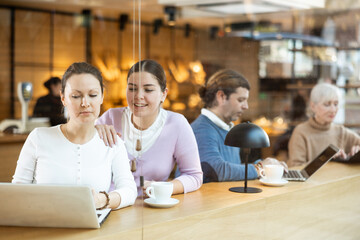 Two interested young female colleagues working on laptop in cafe, sitting at table over cup of coffee, browsing emails and talking