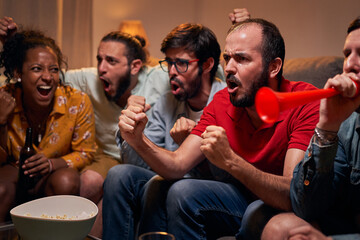 Excited group of male fan screaming and watching match and celebrating goal of favorite team with friends in evening at home. Soccer supporters having fun together sitting on a sofa in the living room - obrazy, fototapety, plakaty