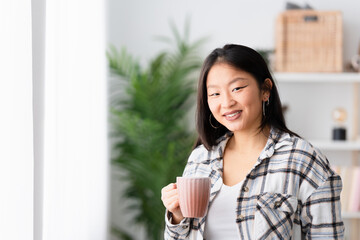 Chinese happy woman drinking coffee at home standing next to a window