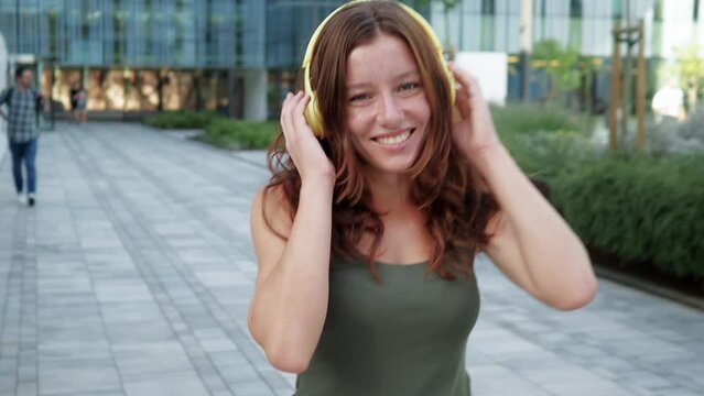 Happy redhead teenage girl with headphones walking in a city and looking at camera. Empowered young woman