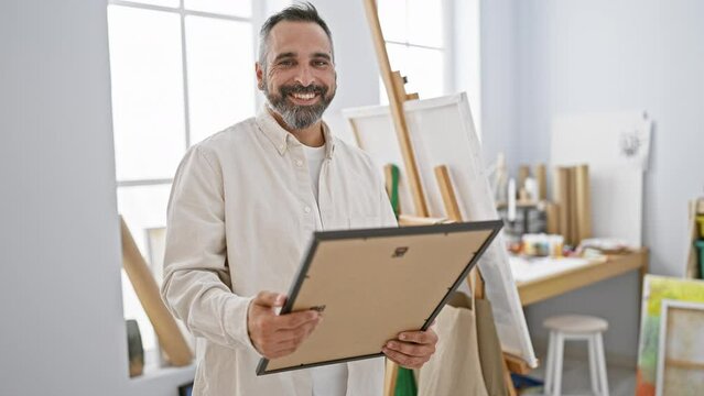 A bearded man with grey hair smiles while holding a clipboard in an art studio full of canvases and painting supplies.