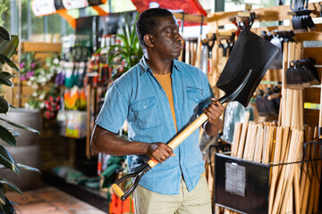 Focused adult african american man choosing shovel to working in his garden at gardening tools store.. - obrazy, fototapety, plakaty