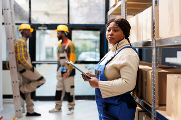 African american warehouse woman operator holding clipboard and standing near carboard boxes shelf....