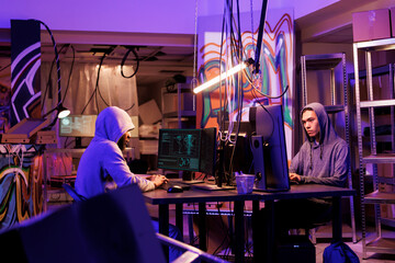 Naklejka premium Internet criminals working together to break into database in dark room. African american and asian young men hacking computer system and installing virus malicious software